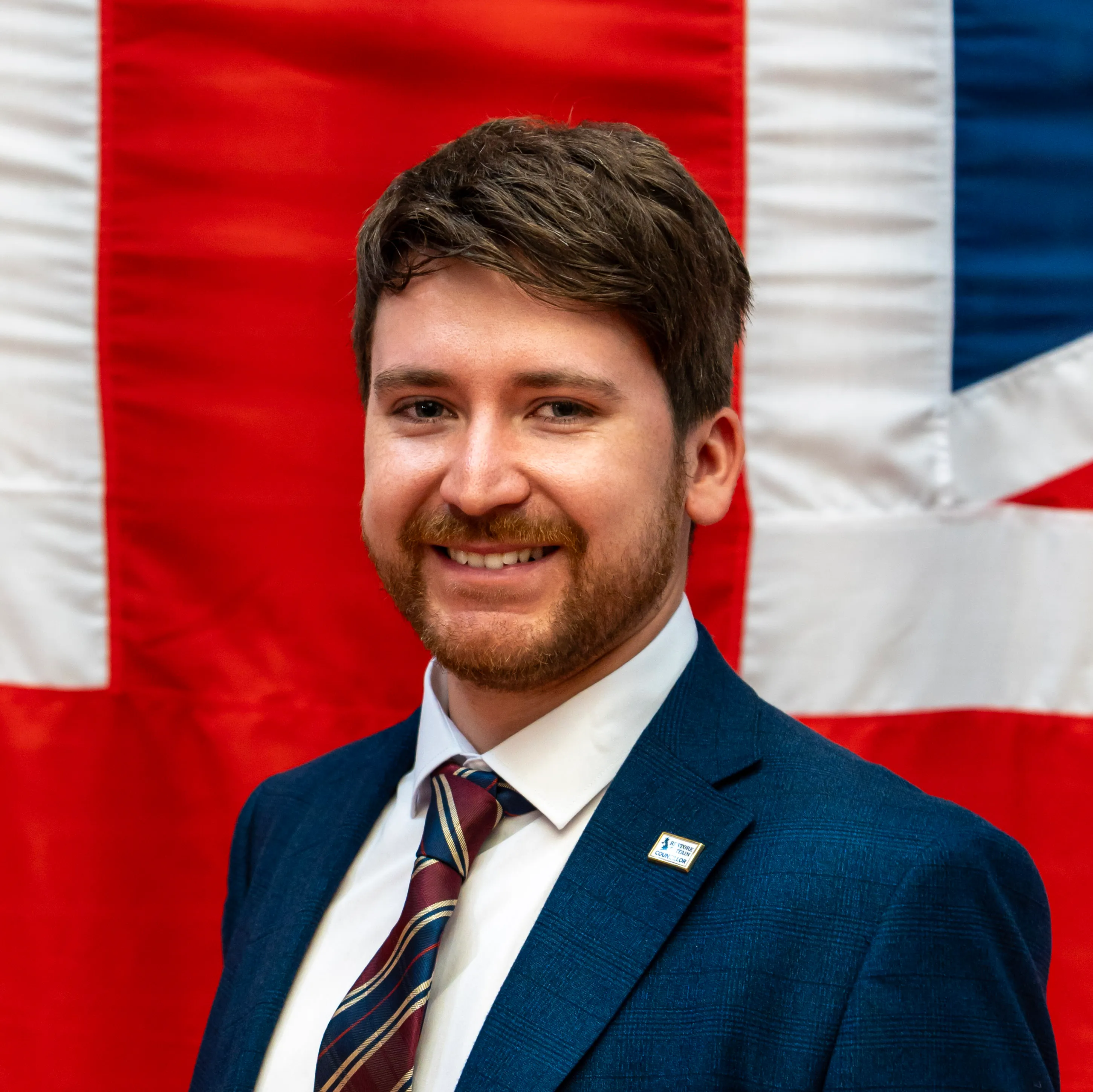 Professional headshot of a smiling man in a navy suit and striped tie against a soft Union Jack backdrop